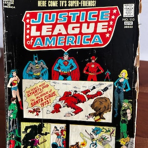 Op de afbeelding: Een vintage Justice League of America stripboek, nummer 110, met een zwarte cover en illustraties van diverse superhelden. De centrale afbeelding toont een gevallen Kerstman met de tekst, The Murder of Santa Claus, 1973.
