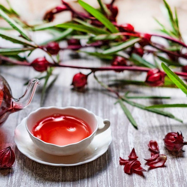 May include: A white teacup and saucer filled with a vibrant red liquid, likely tea, sits on a wooden surface. A glass teapot and sprigs of red berries and green leaves are in the background. The scene suggests a refreshing beverage.