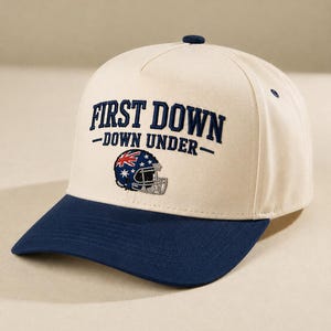May include: A cream-colored baseball cap with a dark blue brim and a dark blue button on top. The front features embroidered dark blue text that reads FIRST DOWN -DOWN UNDER- above an embroidered football helmet with an Australian flag pattern.