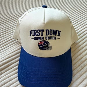 May include: A white and blue baseball cap with the text "FIRST DOWN - DOWN UNDER" in navy blue. The cap features a football helmet graphic with the Australian flag. The brim is blue.