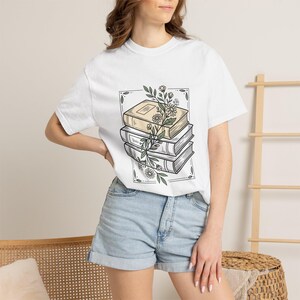 May include: White t-shirt featuring a stack of three books with a floral design. The books are beige and white, with green leaves and white flowers. The design is framed by a rectangular border.
