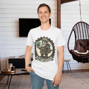 May include: White t-shirt featuring a cartoon gnome artificer design. The illustration shows a bearded gnome holding a glowing orb and tools, set within a gear-shaped frame. The text "Gnome Artificer" is printed around the frame.
