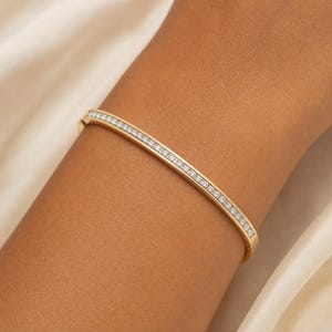 May include: A gold bangle bracelet featuring a continuous row of small, clear, round-cut stones set along the center of the band. The metal has a polished finish, and the design is a slim, circular hoop that encircles the wrist with a secure clasp.