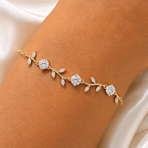 Puede incluir: Una pulsera de cadena dorada con tres piedras redondas de talla brillante y varias piedras de talla marquesa transparentes dispuestas en un patrón de enredadera. Los eslabones metálicos conectan las piedras formando una banda flexible.