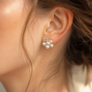 May include: A cluster earring featuring five small, round, cream-coloured pearls arranged in a floral-like pattern. The pearls are set against a silver-toned metal base, which includes a small, delicate silver butterfly accent at the top of the cluster.