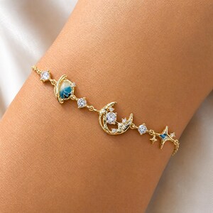 May include: A gold-tone chain bracelet featuring celestial charms including a blue and white enamel planet, a crescent moon with a central clear stone, and a four-pointed star with blue enamel accents, interspersed with small clear round stones.