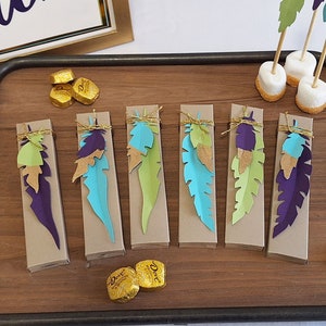 May include: A wooden tray with eight small boxes decorated with colorful feathers. The boxes are tied with twine and have a gold accent. The tray is on a white surface with a sign that says "Wild One" in the background.