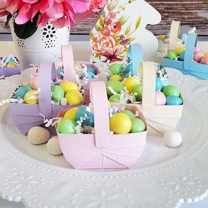 May include: A white plate with a lace design holds several small pastel-colored paper baskets filled with pastel-colored chocolate candies. The baskets have handles and are arranged in a circle on the plate. A bouquet of pink flowers in a white pot is in the background.