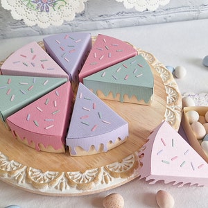 May include: A set of six pastel-colored cake slice boxes with sprinkles and frosting designs. The boxes are arranged in a circle on a wooden cake stand.