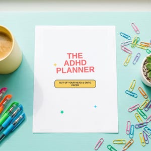 Peut inclure: Vue aérienne d'un espace de travail avec une surface bleu clair. Un planificateur blanc avec le texte "The ADHD Planner" est au centre. Une tasse jaune avec du café, des stylos colorés, des trombones et une plante grasse en pot blanc sont également visibles.