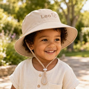 Op de afbeelding: Een beige bucket hat met de geborduurde naam Leo en een klein icoontje van een berengezicht aan de voorzijde. De hoed is voorzien van een bijpassende kinband met een verstelbaar wit koordstopper voor een goede pasvorm en bescherming.