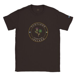 Puede incluir: Camiseta marrón oscuro con un diseño circular con las palabras "SCOTLAND" e "IRELAND" en dorado. El diseño incluye un cardo y un trébol, con el texto "An dá thaigh, aon chroíde". Una pequeña "X" azul en la manga.