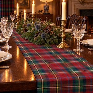 May include: A long rectangular table runner featuring a red, green, blue, and yellow plaid pattern. The runner is centered on a dark wood dining table set with white plates, crystal glasses, brass candlesticks, and a centerpiece of greenery.