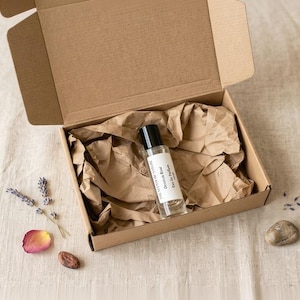 May include: A clear glass rollerball perfume bottle with a black cap and a white label reading "Obsidian Rose Eau De Parfum" nestled inside an open brown cardboard box lined with crumpled brown paper, accompanied by lavender and a rose petal.