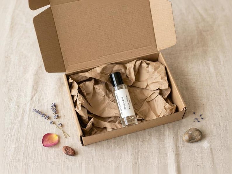 May include: A clear glass rollerball perfume bottle with a black cap and a white label reading "Obsidian Rose Eau De Parfum" nestled inside an open brown cardboard box lined with crumpled brown paper, accompanied by lavender and a rose petal.