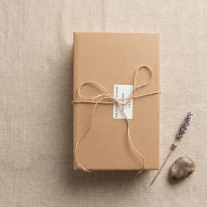 May include: A rectangular cardboard box wrapped in thin brown twine tied in a bow. A white rectangular label is affixed to the center with the text THE SCENT OF NATURE, Eau de Parfum, 3 x 5. A sprig of dried lavender and a smooth brown stone.