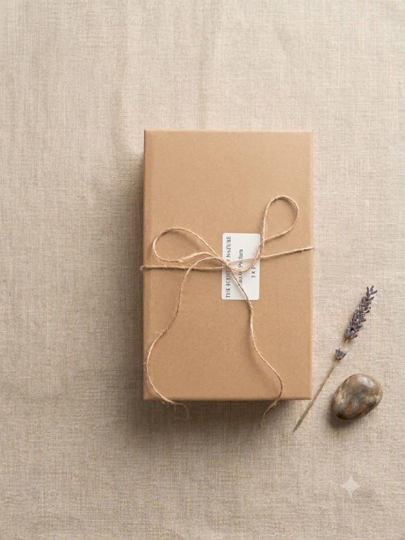 May include: A rectangular cardboard box wrapped in thin brown twine tied in a bow. A white rectangular label is affixed to the center with the text THE SCENT OF NATURE, Eau de Parfum, 3 x 5. A sprig of dried lavender and a smooth brown stone.