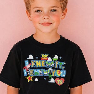 Pode incluir: Uma t-shirt preta de manga curta com um gr&aacute;fico colorido do Toy Story. O design inclui o texto 'I KNEW IT, I KNEW YOU' com padr&otilde;es de personagens, um foguet&atilde;o, uma estrela amarela, nuvens brancas e um cora&ccedil;&atilde;o vermelho com 'TS'.