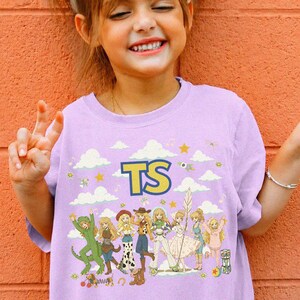 May include: A light purple t-shirt featuring a graphic print of various cartoon characters in a group, with the letters TS in yellow and blue at the top. The design includes white clouds, yellow stars, and small musical notes scattered across the front.