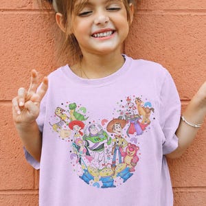 Può includere: Una t-shirt a maniche corte color viola chiaro con una stampa grafica di vari personaggi animati disposti a forma di silhouette di testa di topo. Il design include figure come un ranger spaziale, un cowboy, una cowgirl e diversi piccoli alieni verdi.
