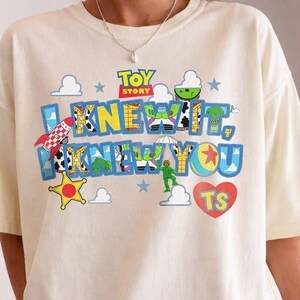 May include: A cream-colored short-sleeve t-shirt featuring a colorful Toy Story graphic. The design includes the text I KNEW IT, I KNEW YOU in blue letters filled with character patterns, a red rocket, a yellow star, clouds, and a red heart with TS.