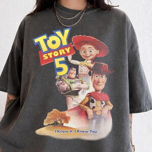 May include: A charcoal gray short-sleeve t-shirt featuring a graphic print of Toy Story 5 with characters Jessie, Buzz Lightyear, and Woody. The design includes the text Toy Story 5 and the phrase I Knew It, I Knew You above a small landscape.