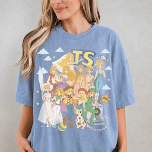 May include: A blue short-sleeve t-shirt featuring a graphic print of multiple blonde cartoon characters in various outfits. The text TS is printed in large yellow letters at the top. The design includes small white clouds, stars, and a rocket.
