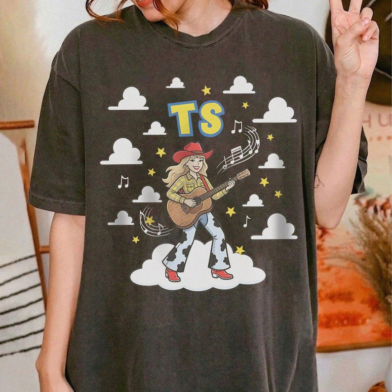 May include: A charcoal grey short-sleeve t-shirt featuring a graphic of a person in a cowboy hat and boots playing an acoustic guitar. The design includes the letters TS, white clouds, yellow stars, and musical notes on a dark background.