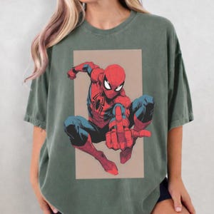 Puede incluir: Una camiseta de manga corta en color verde oliva apagado con un estampado gr&aacute;fico del personaje de c&oacute;mic Spider-Man en posici&oacute;n agachada. El personaje viste un traje rojo y azul con un emblema de ara&ntilde;a negra en el pecho, sobre un fondo rectangular de color beige.