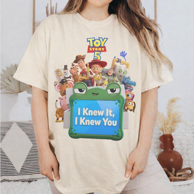 May include: A cream-colored t-shirt featuring a Toy Story 5 graphic with various characters including Woody, Jessie, and Buzz Lightyear. Below the characters is a green frog-shaped frame containing the text I Knew It, I Knew You in blue.
