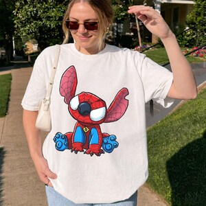 May include: A white short-sleeved t-shirt featuring a graphic print of a blue and red character with large ears and a spider-web pattern on its head and body. The character has large white eyes, blue patches on its chest and feet, and a small spider emblem.