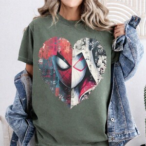 May include: An olive green t-shirt featuring a large heart-shaped graphic on the chest. The graphic is split into two halves, showing a red and black spider-themed mask on the left and a white and black comic book style illustration on the right.