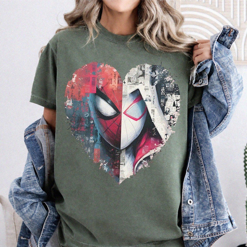 May include: An olive green t-shirt featuring a large heart-shaped graphic on the chest. The graphic is split into two halves, showing a red and black spider-themed mask on the left and a white and black comic book style illustration on the right.
