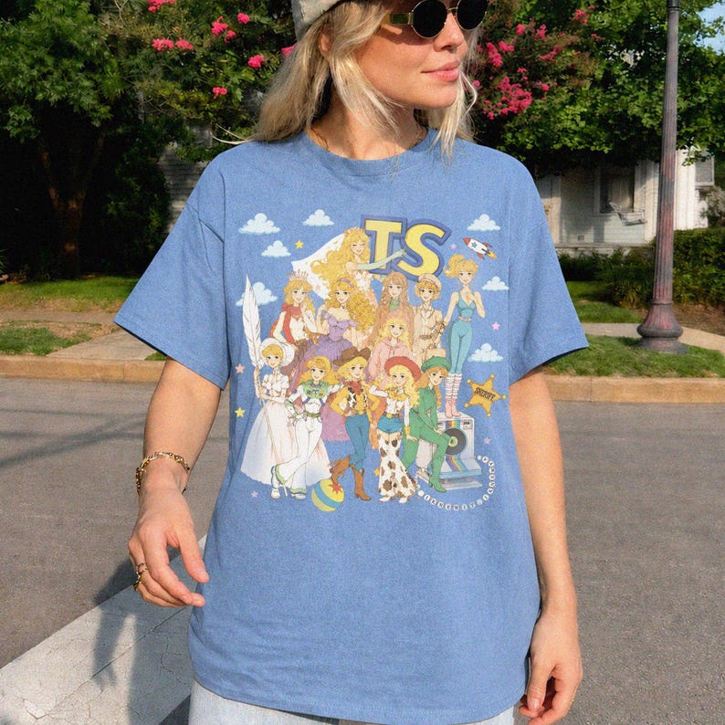 May include: A light blue short-sleeve t-shirt featuring a graphic print of multiple blonde characters in various outfits, including cowgirl attire and dresses. The design includes the letters TS, a rocket, clouds, and a star-shaped badge.
