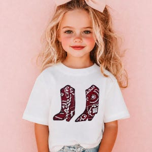 K&ouml;nnte beinhalten: Ein wei&szlig;es kurz&auml;rmeliges T-Shirt mit einem Grafikdruck von zwei dunkelroten Cowboystiefeln, die mit komplizierten wei&szlig;en Blumen- und Wirbelmustern verziert sind. Die Stiefel sind nebeneinander in der Mitte des T-Shirts platziert.