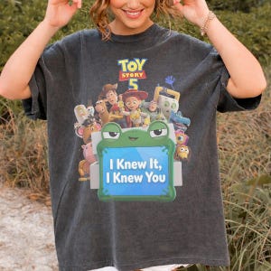 May include: A charcoal gray short-sleeve t-shirt featuring a Toy Story 5 graphic print. The design includes various animated characters and a central green frog-shaped frame containing the text I Knew It, I Knew You in white lettering.