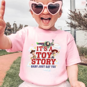 K&ouml;nnte beinhalten: Ein hellrosa T-Shirt mit kurzen &Auml;rmeln und einem Grafikdruck von Woody, Buzz Lightyear und Jessie aus Toy Story. Der Text lautet IT'S A TOY STORY BABY JUST SAY YES in roter und blauer Schrift, umgeben von wei&szlig;en Wolken und gelben Sternen.