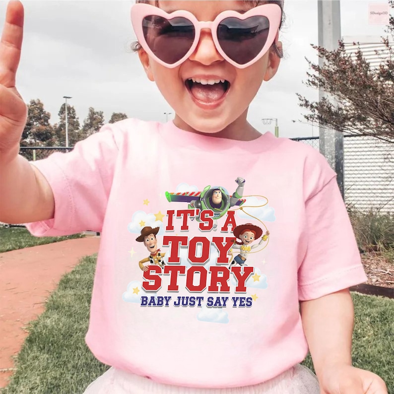 K&ouml;nnte beinhalten: Ein hellrosa T-Shirt mit kurzen &Auml;rmeln und einem Grafikdruck von Woody, Buzz Lightyear und Jessie aus Toy Story. Der Text lautet IT'S A TOY STORY BABY JUST SAY YES in roter und blauer Schrift, umgeben von wei&szlig;en Wolken und gelben Sternen.