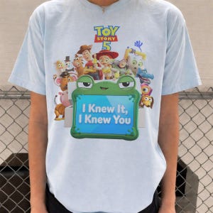May include: A light blue t-shirt featuring a Toy Story 5 graphic with various characters and a green frog-shaped frame containing the text I Knew It, I Knew You. The design includes Woody, Jessie, Buzz Lightyear, and other animated figures.