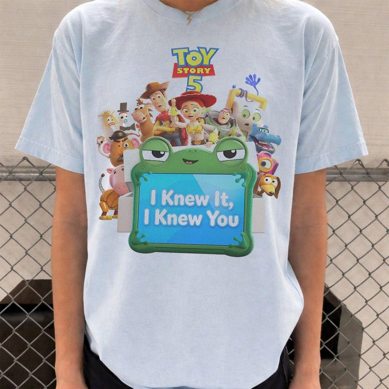 May include: A light blue t-shirt featuring a Toy Story 5 graphic with various characters and a green frog-shaped frame containing the text I Knew It, I Knew You. The design includes Woody, Jessie, Buzz Lightyear, and other animated figures.
