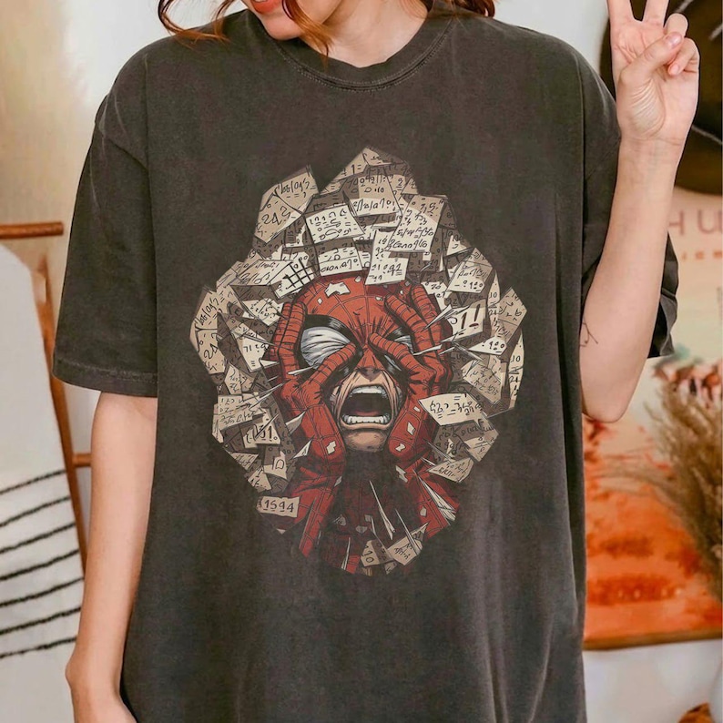 May include: A charcoal gray short-sleeve t-shirt featuring a graphic print of a red masked character with hands pressed against the face, surrounded by a collage of scattered, overlapping paper fragments covered in handwritten black ink notes.