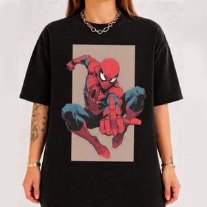 Puede incluir: Una camiseta negra de manga corta con un estampado gr&aacute;fico de un superh&eacute;roe rojo y azul en posici&oacute;n agachada. El personaje tiene un emblema de ara&ntilde;a en el pecho y extiende un dedo hacia el espectador. El dise&ntilde;o est&aacute; dentro de un rect&aacute;ngulo.