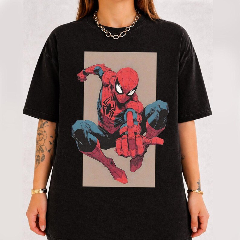 Puede incluir: Una camiseta negra de manga corta con un estampado gr&aacute;fico de un superh&eacute;roe rojo y azul en posici&oacute;n agachada. El personaje tiene un emblema de ara&ntilde;a en el pecho y extiende un dedo hacia el espectador. El dise&ntilde;o est&aacute; dentro de un rect&aacute;ngulo.