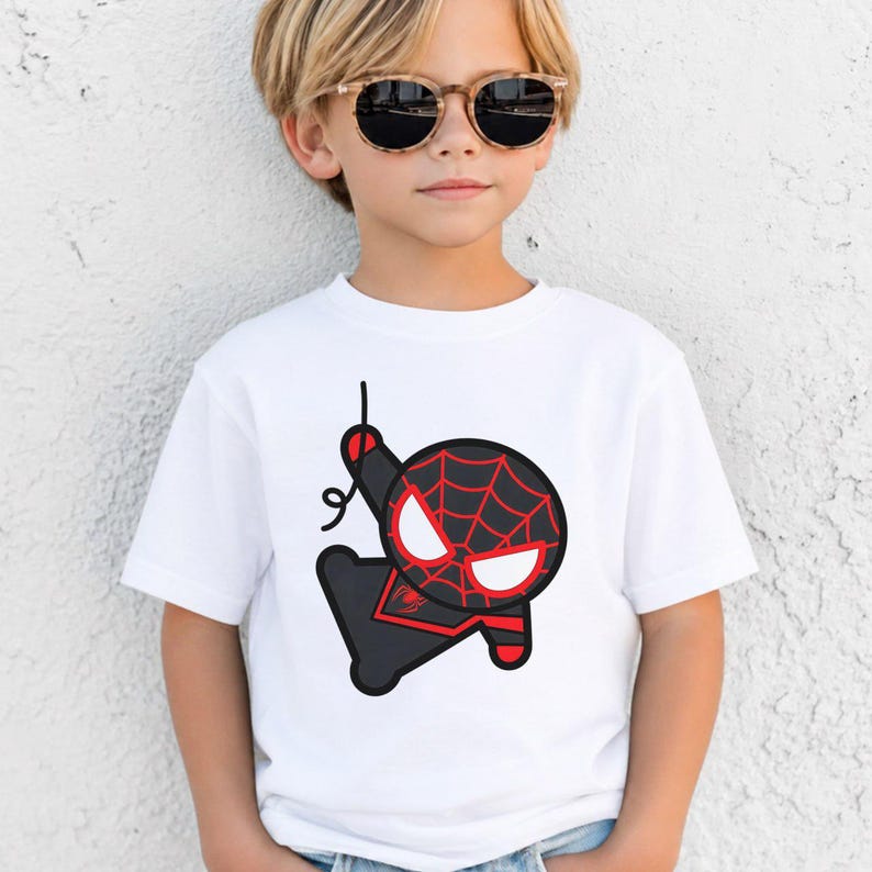 May include: A white short-sleeved t-shirt featuring a graphic print of a stylised, cartoon-like character with a black and red spider-web pattern mask, large white eyes, and a black suit, shown hanging from a thin black line on the chest.