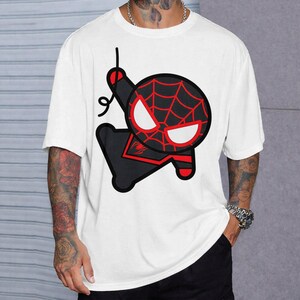 May include: A white short-sleeved t-shirt featuring a large, stylised graphic of a cartoon character with a black and red spiderweb mask, large white eyes, and a black suit with red accents, depicted in a swinging pose with a black web line.