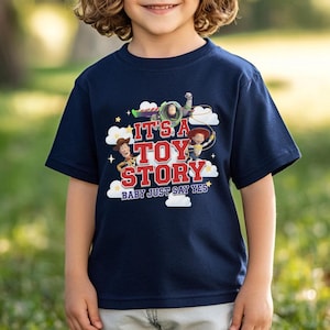 K&ouml;nnte beinhalten: Ein marineblaues Kurzarm-T-Shirt mit einem Grafikdruck von Woody, Buzz Lightyear und Jessie aus Toy Story. Der Text lautet IT'S A TOY STORY BABY JUST SAY YES, umgeben von wei&szlig;en Wolken und gelben Sternen auf der Vorderseite des Kleidungsst&uuml;cks.