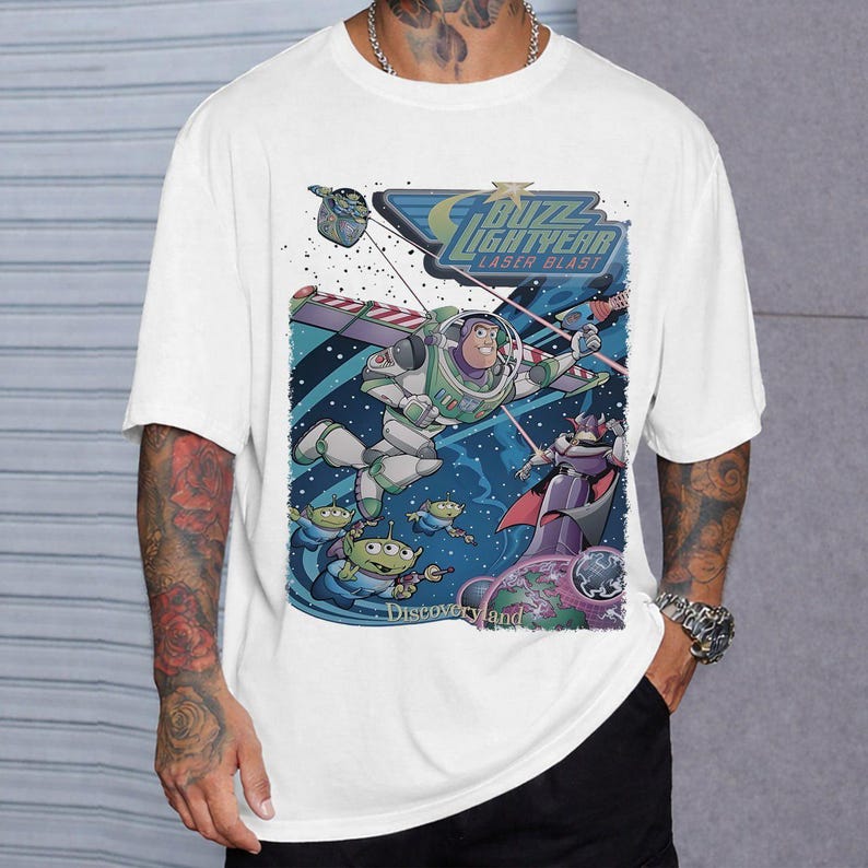 May include: White t-shirt featuring a colorful graphic of Buzz Lightyear in a space battle scene. The design includes the text "Buzz Lightyear Laser Blast" and "Discoveryland". The artwork showcases characters from the Toy Story franchise.