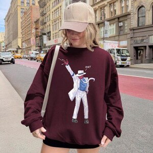 May include: A maroon long-sleeve sweatshirt featuring a graphic print of a figure in a white suit, blue shirt, and white fedora, striking a dance pose with one hand raised and the text AOW!! printed in white near the figure's head.