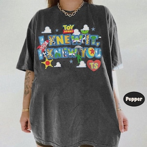 Pode incluir: Uma t-shirt de manga curta cinzento-carv&atilde;o com uma impress&atilde;o gr&aacute;fica colorida com o texto TOY STORY I KNEW YOU TS. O design inclui v&aacute;rios padr&otilde;es tem&aacute;ticos de personagens, um foguet&atilde;o vermelho, uma estrela amarela e um cora&ccedil;&atilde;o vermelho.