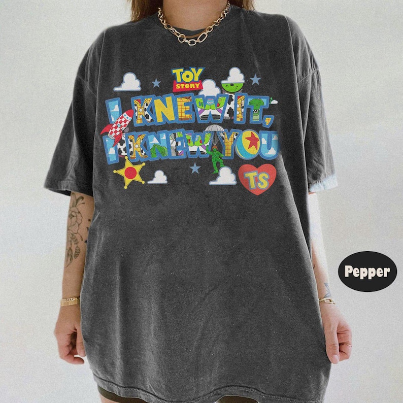 Pode incluir: Uma t-shirt de manga curta cinzento-carv&atilde;o com uma impress&atilde;o gr&aacute;fica colorida com o texto TOY STORY I KNEW YOU TS. O design inclui v&aacute;rios padr&otilde;es tem&aacute;ticos de personagens, um foguet&atilde;o vermelho, uma estrela amarela e um cora&ccedil;&atilde;o vermelho.